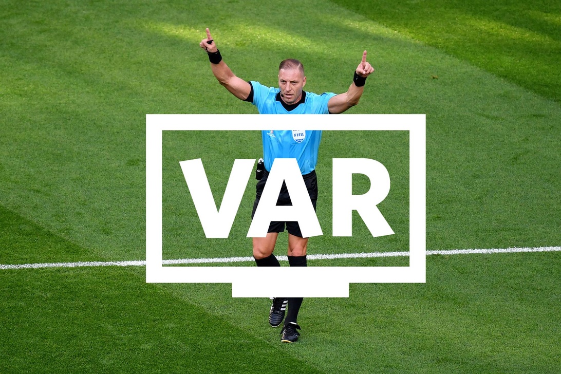 Jalkapallotuomari sinisessä FIFA-asussa tekemässä VAR-merkkiä vihreällä kentällä, VAR-logo kuvan päällä.